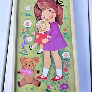 May include: A framed illustration of a girl holding a doll in a garden setting. The girl is wearing a purple dress and has brown hair with a black bow. The doll is wearing a red dress and has blonde hair. A brown teddy bear with a pink bow is sitting in the grass. The illustration is signed "FK".