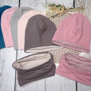May include: A collection of soft, solid-colored beanies and neck gaiters in various shades, including teal, navy, pink, gray, and brown. The beanies are folded and displayed alongside matching neck warmers, showcasing a coordinated set of accessories.