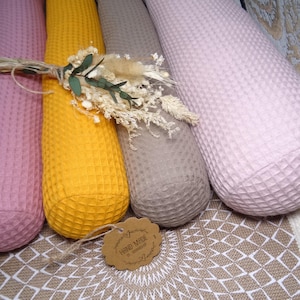 Könnte beinhalten: Vier zylindrische, strukturierte Stoffartikel in Rosa, Gelb, Grau und Hellrosa werden gezeigt. Ein Bündel Trockenblumen und ein Etikett mit der Aufschrift „Hand Made“ sind oben platziert. Die Artikel sind auf einer gemusterten Oberfläche angeordnet.
