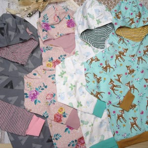 Könnte beinhalten: Eine Auswahl an Kinder-Hoodies mit verschiedenen Mustern. Designs umfassen Blumen, Rehe und geometrische Formen. Die Hoodies haben kontrastierende Bündchen und Kapuzenfutter in Farben wie Rosa, Grau und Türkis. Der Stoff scheint weich und bequem zu sein.