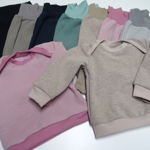 Könnte beinhalten: Eine Sammlung von gerippten Strickpullovern in verschiedenen Farben, darunter rosa, beige, grau, grün und schwarz. Die Pullover haben einen Rundhalsausschnitt und lange Ärmel.