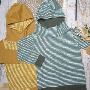 Könnte beinhalten: Zwei Kapuzenpullover. Einer ist senfgelb mit weißen Streifen, der andere ist türkis mit weißen horizontalen Streifen. Beide haben eine lockere Passform und eine Kapuze. Die Sweatshirts sind aus einem weichen Material.