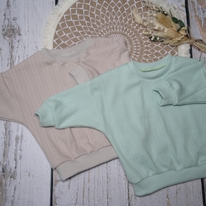Könnte beinhalten: Zwei pastellfarbene Sweatshirts aus Rippstrick. Eines ist zartrosa, das andere hellmintgrün. Beide haben einen Rundhalsausschnitt und lange Ärmel mit lockerer Passform. Die Sweatshirts werden auf einer hellen Holzoberfläche präsentiert.