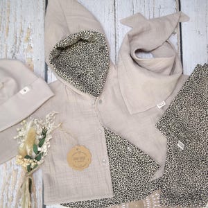 Könnte beinhalten: Ein Set Babykleidung in neutralem Beige mit Leopardenmuster. Das Set beinhaltet eine Kapuzenjacke, ein Lätzchen, eine Mütze und ein Spucktuch.