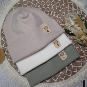 Könnte beinhalten: Drei Strickmützen in verschiedenen Farben: hellrosa, weiß und grün. Jede Mütze hat ein kleines Leder-Etikett mit einem Logo. Die Mützen sind übereinander gestapelt.