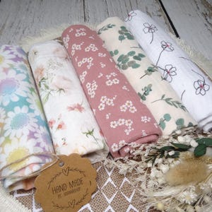 Könnte beinhalten: Fünf handgefertigte Musselin-Swaddles mit Blumenmustern. Die Swaddles sind gefaltet und auf einer gewebten Oberfläche angeordnet. Ein Etikett mit dem Text "Hand Made by Woodnwick" ist an den Swaddles befestigt.