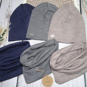Könnte beinhalten: Eine Auswahl an Mützen und Halstüchern in Marineblau, Grau und Beige. Die Mützen haben ein schlichtes Design, während die Halstücher gefaltet sind. Die Artikel sind aus einem weichen Strickstoff gefertigt. Ein kleines Etikett ist sichtbar.