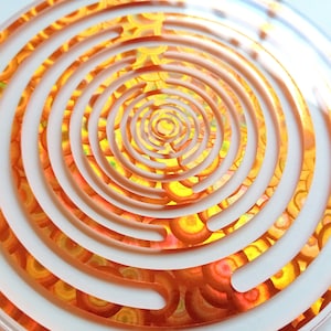 Op de afbeelding: Een helder, cirkelvormig decoratief stuk met een spiraalvormig ontwerp. Het ontwerp heeft concentrische ringen met een uitgesneden patroon, dat een kleurrijke, patroonachtergrond in tinten oranje, geel en rood onthult. Het stuk is waarschijnlijk gemaakt van acryl.