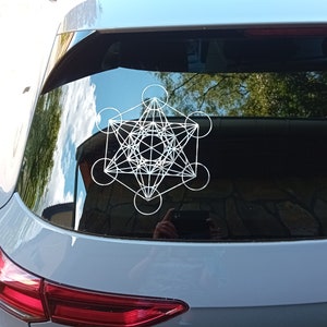 Puede incluir: Una pegatina de diseño geométrico blanco en la ventana trasera de un coche gris. El diseño es un patrón complejo de líneas y círculos, que se asemeja a un símbolo de geometría sagrada.