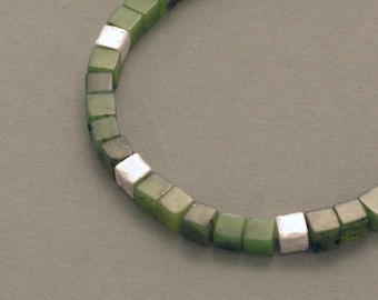 Op de afbeelding: Een groene en zilveren armband gemaakt van vierkante kralen. De kralen wisselen van kleur, waarbij de groene kralen groter zijn dan de zilveren kralen.