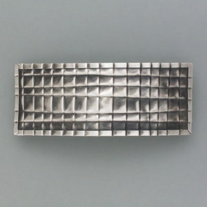 Peut inclure: Un plateau rectangulaire en métal argenté avec un motif texturé en forme de grille. Le plateau a un bord surélevé et une finition mate. Le design est composé de nombreuses petites indentations carrées, créant une surface tactile. Le plateau est probablement utilisé à des fins décoratives ou fonctionnelles.