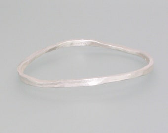 Puede incluir: Un brazalete plateado con textura martillada. El brazalete tiene una forma irregular y orgánica, presentado sobre un fondo gris claro. Esta pieza de joyería es un accesorio sencillo y elegante.