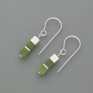 Op de afbeelding: Een paar zilveren oorhangers met groene jade-steentjes. De oorbellen hebben een eenvoudig ontwerp met drie vierkante jade-steentjes aan elke oorbel.