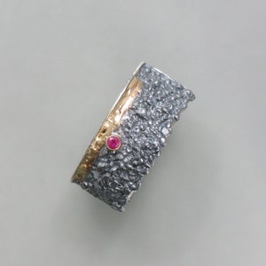 May include: A silver ring with a textured surface and a thin gold band. A small pink gemstone is set in the gold band.