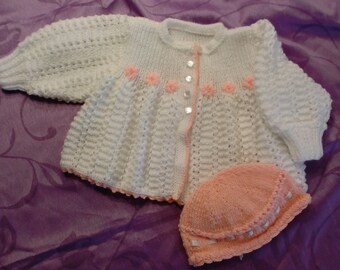 May include: A white knitted baby cardigan with pink flower accents and a matching pink knitted hat.