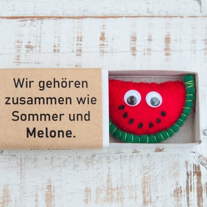 Könnte beinhalten: Eine kleine Streichholzschachtel mit einem braunen Papieretikett, auf dem "Wir gehören zusammen wie Sommer und Melone." steht. Die Schachtel enthält eine Filz-Wassermelone mit Glitzern und einer grünen Filzschale.