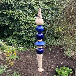 Könnte beinhalten: Ein dekorativer Gartenpfahl mit einem gestapelten Design. Er verfügt über blaue Keramikkugeln, beige steinartige Formen und eine strukturierte, kegelförmige Spitze. Der Pfahl steht in einem Gartenbeet mit grünem Laub im Hintergrund.