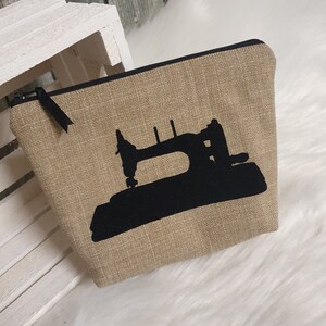 Könnte beinhalten: Eine beige Reißverschlusstasche mit einer schwarzen Silhouette einer Nähmaschine auf der Vorderseite.