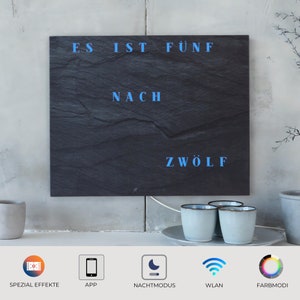Peut inclure: Une horloge en ardoise noire avec un texte bleu illuminé qui indique "Es ist fünf nach zwölf". L'horloge est montée sur un mur blanc. Sous l'horloge, trois tasses en céramique blanche sur une soucoupe blanche.