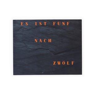 Peut inclure: Une ardoise rectangulaire gris foncé avec le texte orange "ES IST FÜNF NACH ZWÖLF". La surface est texturée, donnant un aspect naturel. Le texte est centré et clairement visible.