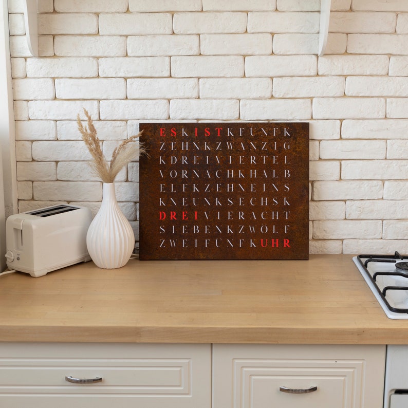 May include: A rusty metal wall art piece with a grid of letters spelling out the German words for numbers one through twelve. The words are highlighted in red. The art piece is hanging on a white brick wall in a kitchen.