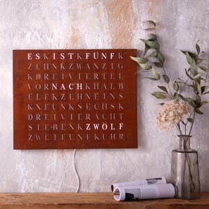 May include: A rusty metal wall clock with white numbers and letters spelling out the German words for numbers. The clock is hanging on a light gray wall with a vase of dried flowers and a magazine on a wooden table below.