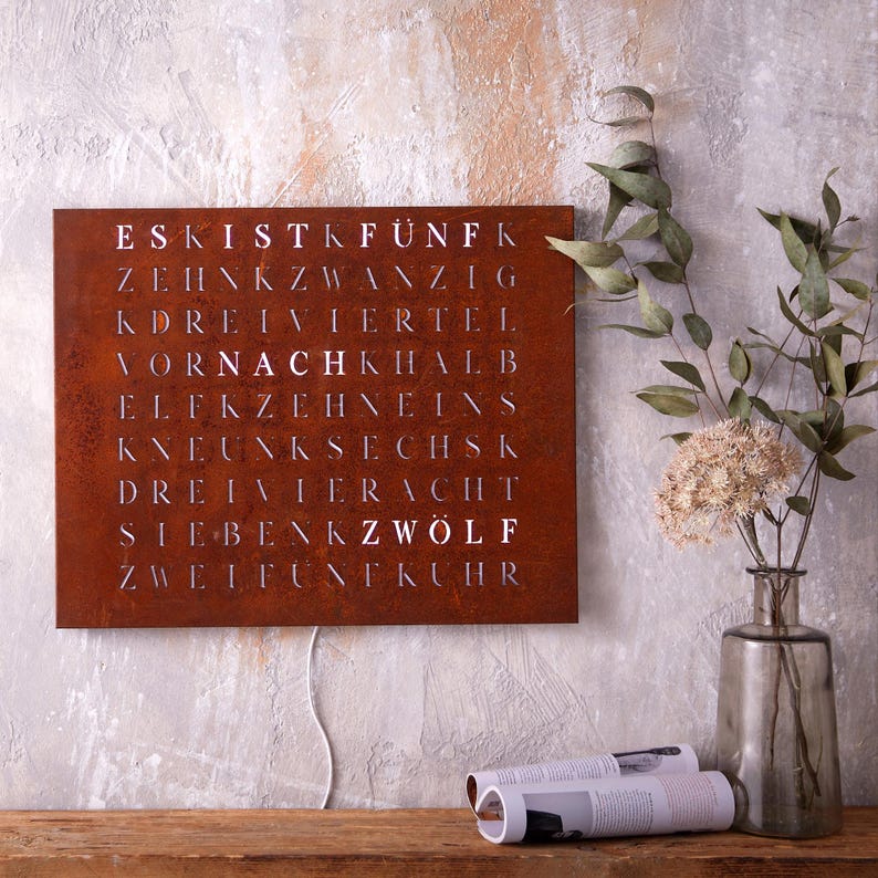 May include: A rusty metal wall clock with white numbers and letters spelling out the German words for numbers. The clock is hanging on a light gray wall with a vase of dried flowers and a magazine on a wooden table below.