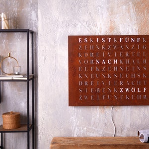 May include: A rusty metal wall sign with white lettering spelling out the German numbers one through twelve. The sign is hanging on a gray wall with a wooden shelf and decorative items in the background.