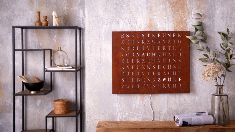 May include: A rusty metal wall sign with white lettering spelling out the German numbers one through twelve. The sign is hanging on a gray wall with a wooden shelf and decorative items in the background.