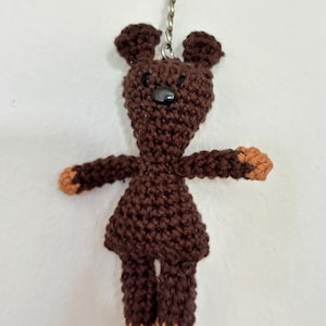 Könnte beinhalten: Ein handgefertigter brauner Häkelbär-Schlüsselanhänger mit einer schwarzen Knopf-Nase und kleinen Ohren. Die Pfoten und Füße des Bären sind hellbraun. Der Schlüsselanhänger ist an einem silbernen Metallring befestigt.