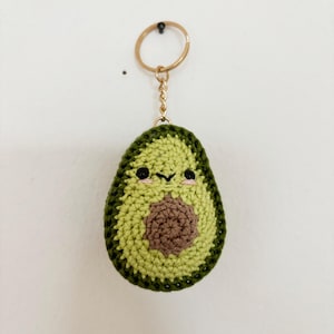 Könnte beinhalten: Ein grüner gehäkelter Avocado-Schlüsselanhänger mit einem braunen Kern und einem niedlichen Gesicht. Der Schlüsselanhänger hat einen goldenen Metallring und eine Kette.
