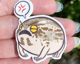 Puede incluir: Una pegatina troquelada que muestra una rana redonda con manchas marrones y beige, expresión facial gruñona, ojos amarillos y boca oscura. Sobre la rana hay un bocadillo blanco con un símbolo rojo de enfado en su interior.