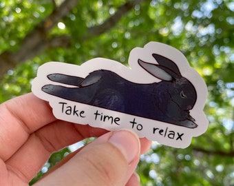 May include: A sticker featuring a black rabbit in a relaxed pose. The sticker has the text "Take time to relax" in a handwritten style font. The background is a blurred view of green foliage.
