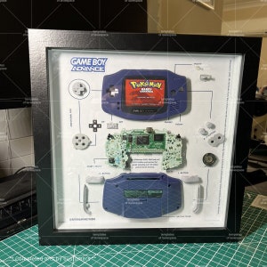 Może przedstawiać: Oprawiony wyświetlacz rozmontowanego Game Boy Advance, pokazujący poszczeg&oacute;lne komponenty przenośnej konsoli do gier. Wyświetlacz zawiera płytę gł&oacute;wną konsoli, przyciski i ekran, a także kartridż gry z tekstem "Pokemon Ruby Version".