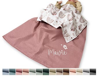 May include: A dusty rose waffle-knit blanket featuring the name Mavie and a small bird graphic in white, partially covered by a white blanket with a repeating brown and tan rainbow pattern, draped over a baby wearing a tan sweater.
