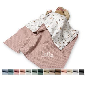 Könnte beinhalten: Eine altrosa Waffelstrick-Babydecke mit dem Namen Lotta in weißer Schreibschrift. Die Decke ist so gefaltet, dass eine weiße Unterseite mit einem Muster aus braunen Rehen, Füchsen und grünen Bäumen sichtbar ist. Ein Baby ist oben zu sehen.