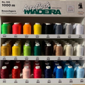 Könnte beinhalten: Eine Präsentation von Aero flock Madera Garnspulen in verschiedenen Farben, darunter Neongrün, Pink, Orange und Gelb. Das Etikett lautet "Nr. 100, 1000 m". Das Garn ist 100 % Polyester und hergestellt in Deutschland.