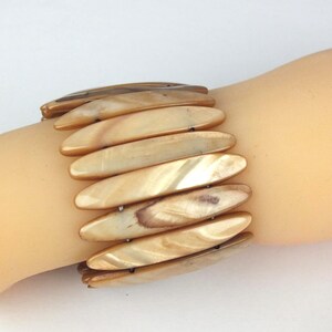 Könnte beinhalten: Ein Stretch-Armband mit länglichen, irisierenden, beigefarbenen Muschelteilen. Das Armband besteht aus mehreren Muschelsegmenten, die durch Gummibänder verbunden sind und eine flexible Passform ermöglichen. Die Muschelteile haben eine natürliche, polierte Oberfläche.