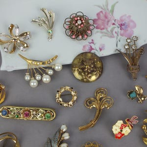 May include: A collection of assorted vintage-style brooches featuring gold-tone metal, faux pearls, clear rhinestones, and colorful enamel accents. The pins include floral, circular, and abstract shapes, some with filigree details and multi-colored stones.