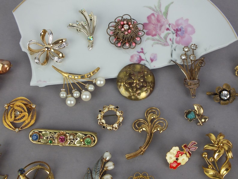 May include: A collection of assorted vintage-style brooches featuring gold-tone metal, faux pearls, clear rhinestones, and colorful enamel accents. The pins include floral, circular, and abstract shapes, some with filigree details and multi-colored stones.
