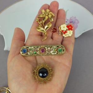May include: A collection of four brooches including a gold-tone flower, a small bouquet with red and white enamel, a rectangular bar pin with blue, pink, and green rhinestones, and a round brooch featuring a central black cabochon stone.