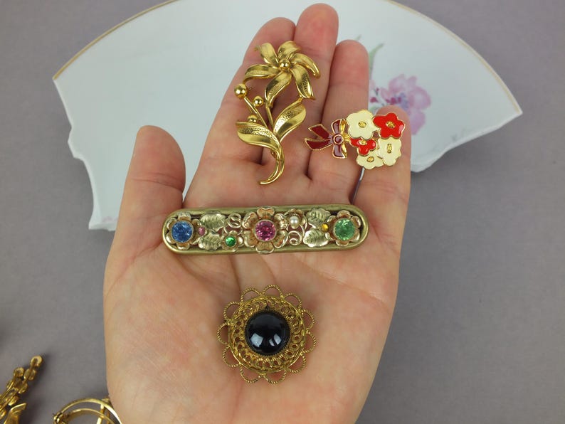 May include: A collection of four brooches including a gold-tone flower, a small bouquet with red and white enamel, a rectangular bar pin with blue, pink, and green rhinestones, and a round brooch featuring a central black cabochon stone.