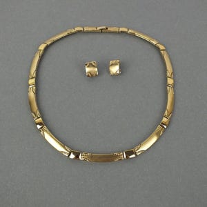 K&ouml;nnte beinhalten: Ein goldfarbenes Halsketten- und Ohrring-Set. Die Halskette ist im Choker-Stil mit einer Reihe von rechteckigen und gebogenen Segmenten. Die Ohrringe sind kleine, quadratische Ohrstecker. Das Schmuckset wird auf grauem Hintergrund pr&auml;sentiert.