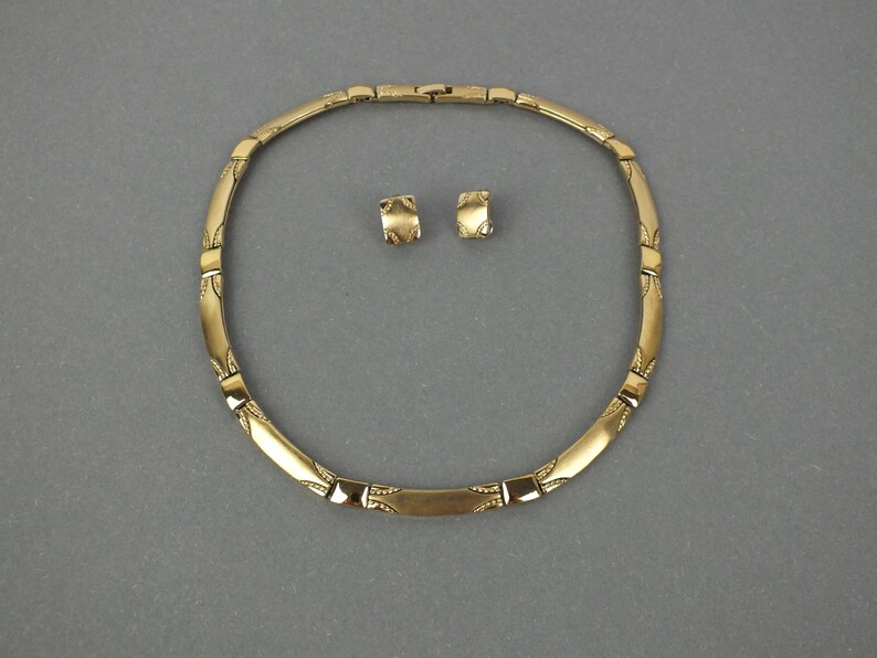 K&ouml;nnte beinhalten: Ein goldfarbenes Halsketten- und Ohrring-Set. Die Halskette ist im Choker-Stil mit einer Reihe von rechteckigen und gebogenen Segmenten. Die Ohrringe sind kleine, quadratische Ohrstecker. Das Schmuckset wird auf grauem Hintergrund pr&auml;sentiert.