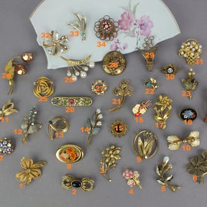May include: A collection of thirty-four assorted vintage-style metal brooches in various shapes including flowers, bows, and abstract designs, featuring gold-tone finishes, colorful enamel accents, faux pearls, and clear rhinestones, all numbered.