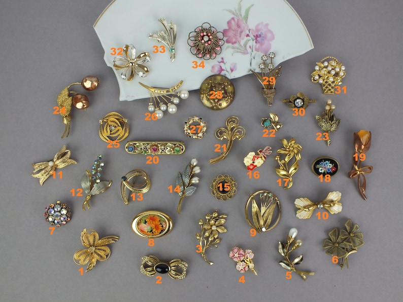 May include: A collection of thirty-four assorted vintage-style metal brooches in various shapes including flowers, bows, and abstract designs, featuring gold-tone finishes, colorful enamel accents, faux pearls, and clear rhinestones, all numbered.