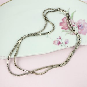 Peut inclure: Un collier argenté avec un motif de chaîne tressée, posé sur une assiette décorative blanche avec des motifs floraux roses. Le collier est présenté sur un fond rose clair, créant une esthétique vintage.