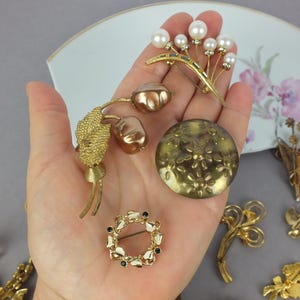 May include: A collection of four gold-tone metal brooches held in a palm. One features white faux pearls, another has two metallic brown buds, one is a round disc with embossed flowers, and the fourth is a small circular wreath with black accents.