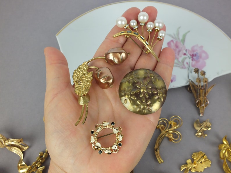 May include: A collection of four gold-tone metal brooches held in a palm. One features white faux pearls, another has two metallic brown buds, one is a round disc with embossed flowers, and the fourth is a small circular wreath with black accents.