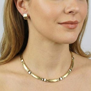 K&ouml;nnte beinhalten: Ein goldfarbenes Halsketten- und Ohrring-Set. Die Halskette ist im Choker-Stil mit gebogenen, segmentierten Gliedern. Die Ohrringe sind quadratisch geformt und haben ein &auml;hnliches Design. Der Schmuck wird von einer Person getragen.